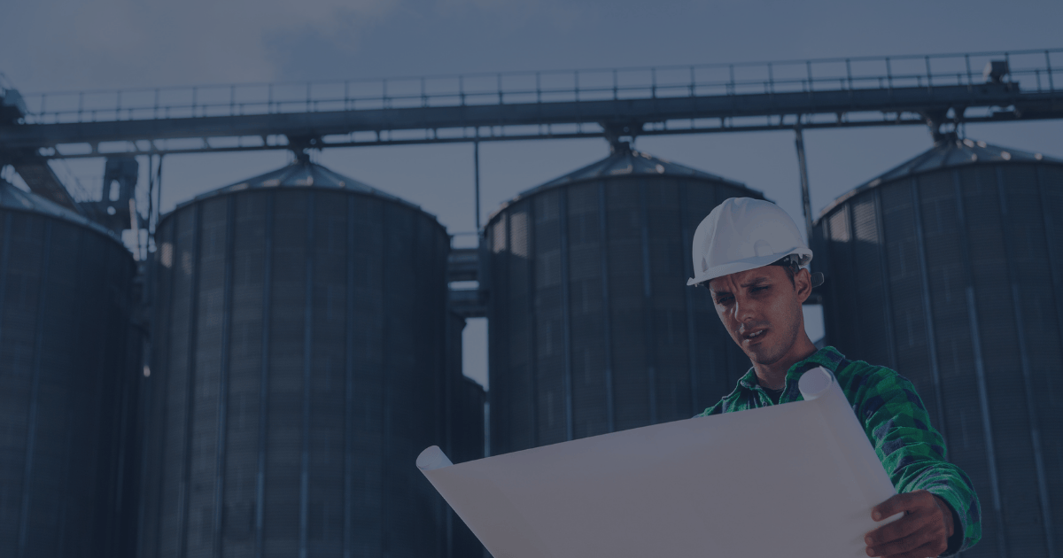An operations manager in a white hard hat reviews a large plant layout blueprint outside an industrial facility with multiple metal silos. The image suggests planning, assessment, and consideration of plant expansion or optimization.        Ask ChatGPT
