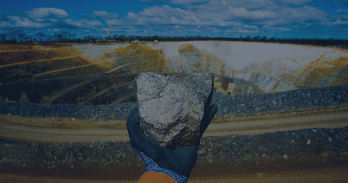 Gloved hand holding a chunk of ore with an open-pit mine in the background, symbolizing the challenge of accurate ore classification from drill to stockpile.
