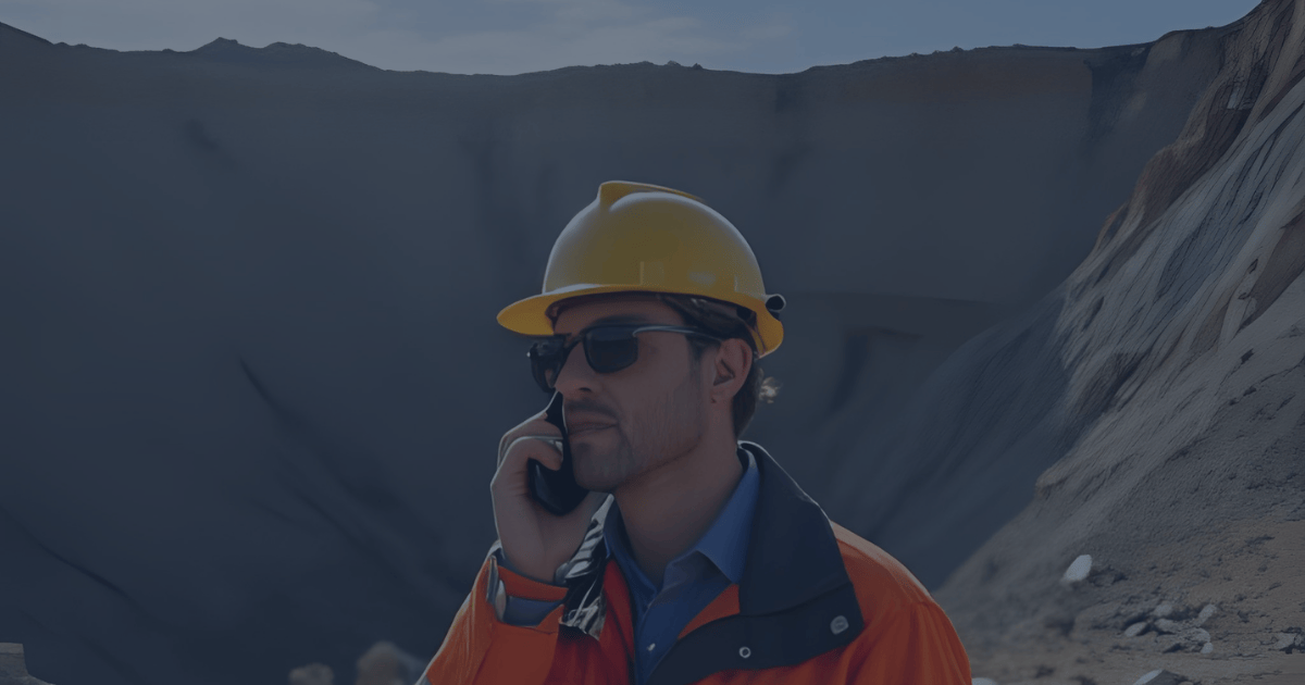 Mining engineer in hard hat speaking on phone in front of a large open-pit mine, symbolizing the communication challenges of coordinating mine-to-mill operations.