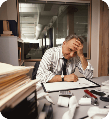 Stressed office worker sitting at a desk with his head in his hand, surrounded by paperwork and files.